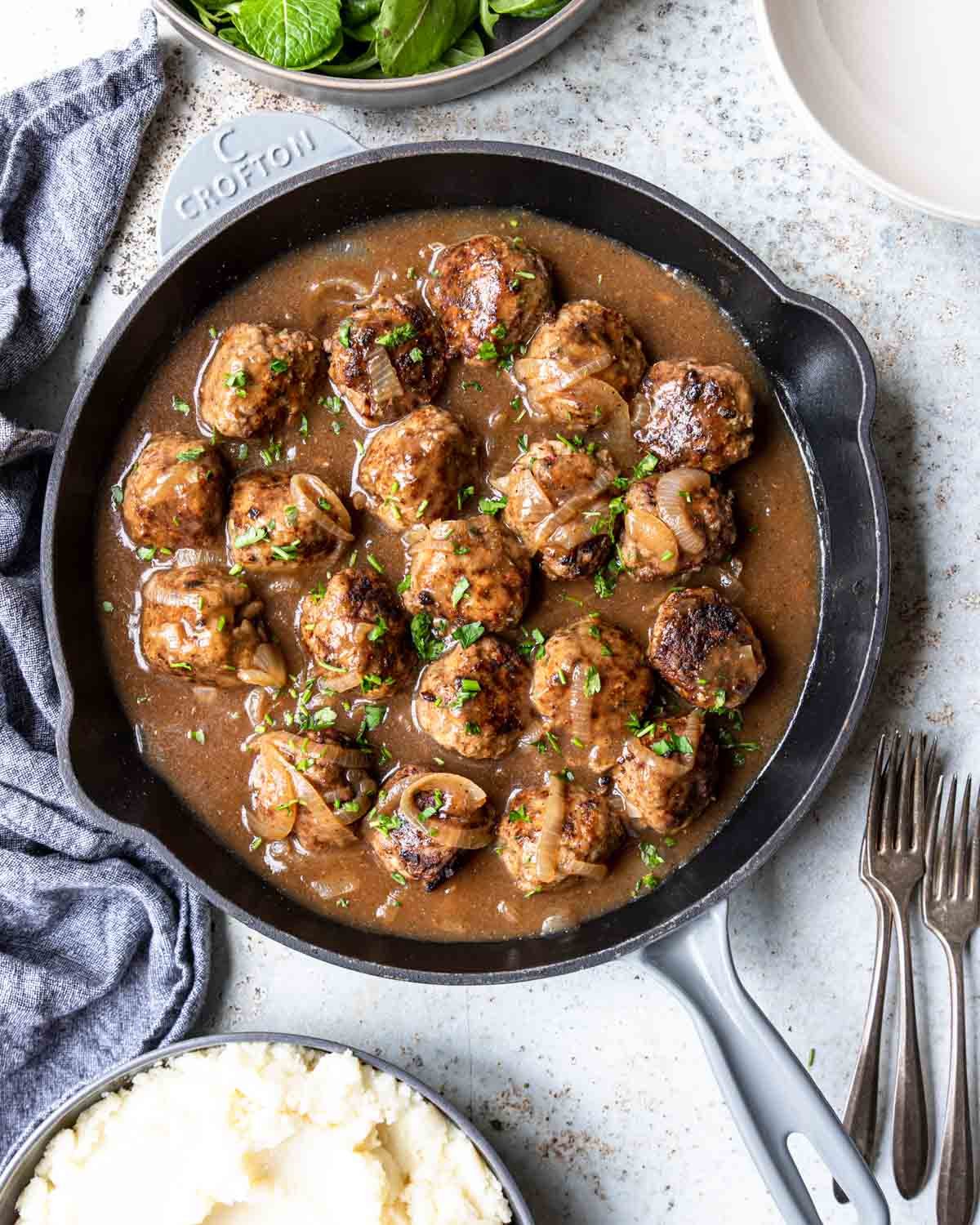 a skillet full of meatballs in a gravy with a salad and mashed potatoes to the side