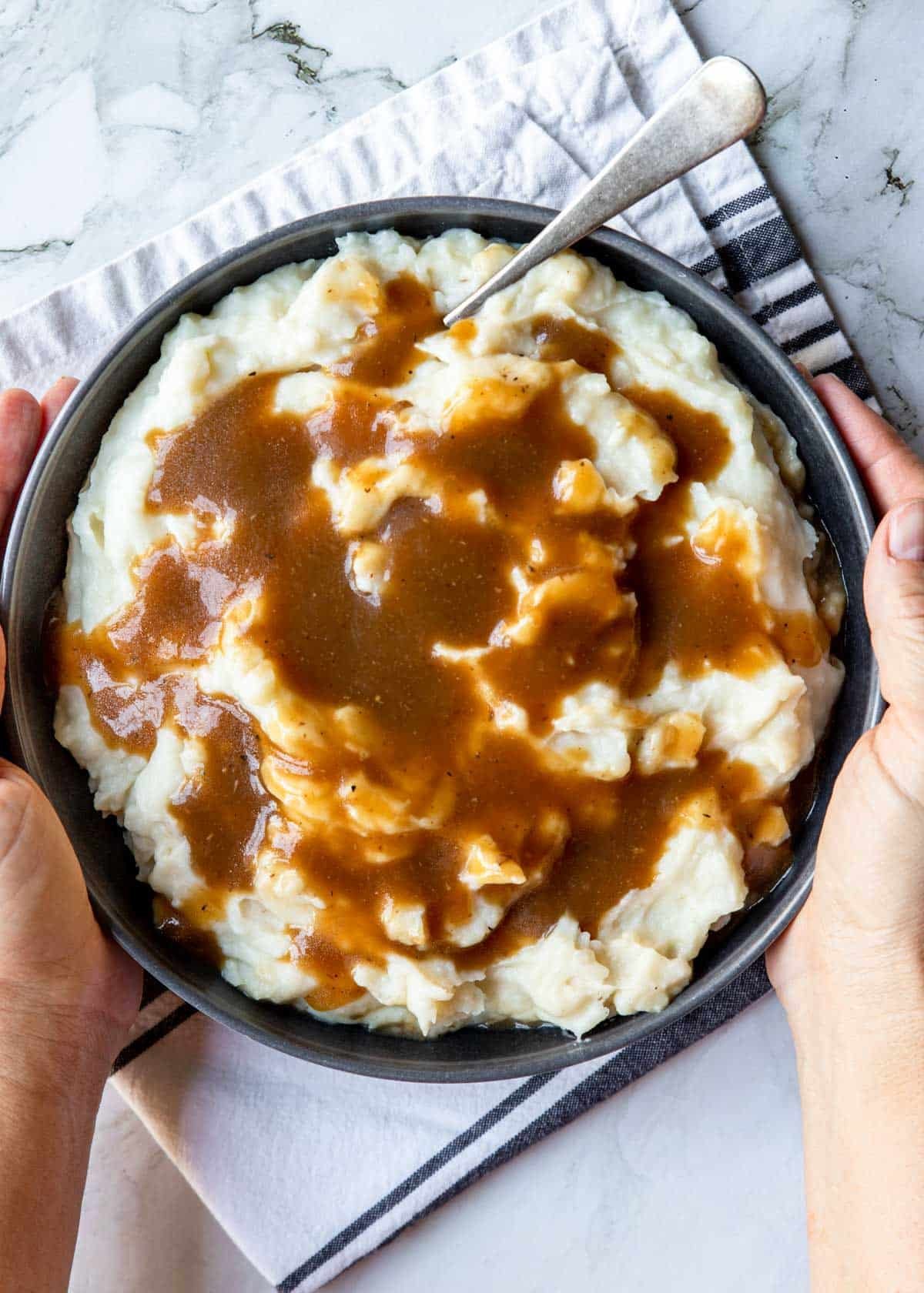 two hands holding a bowl of mashed potatoes with brown gravy poured over top