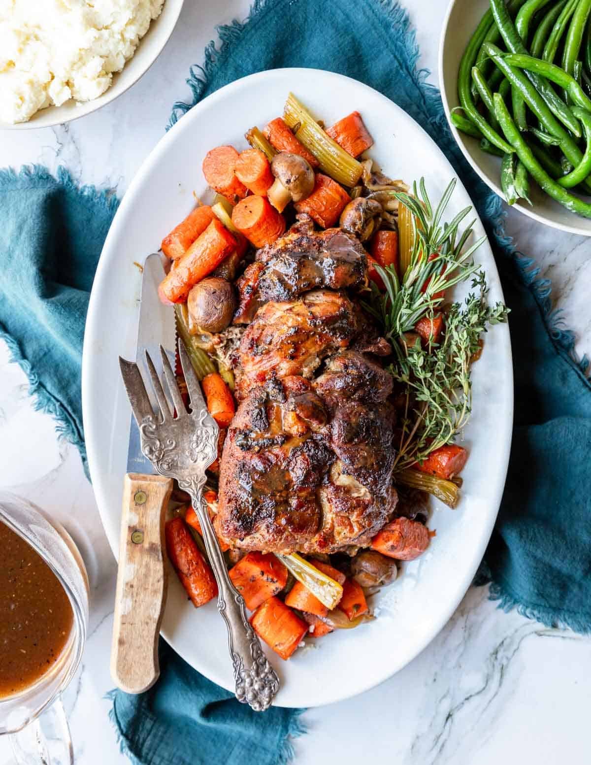 a pork shoulder roast on a plate with cooked carrots, celery, mushrooms and herbs