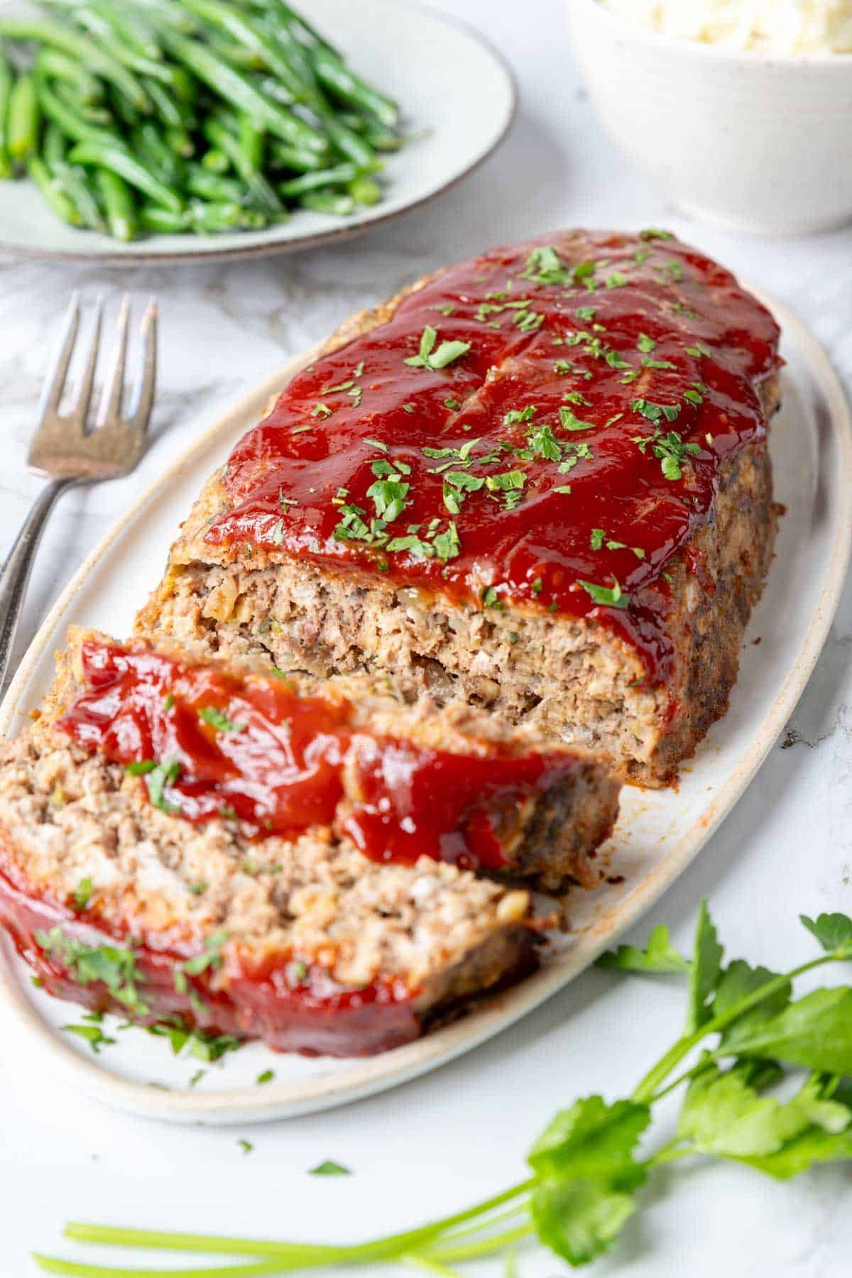 a meatloaf on a platter with two slices cut. green beans and mashed potatoes in the background 