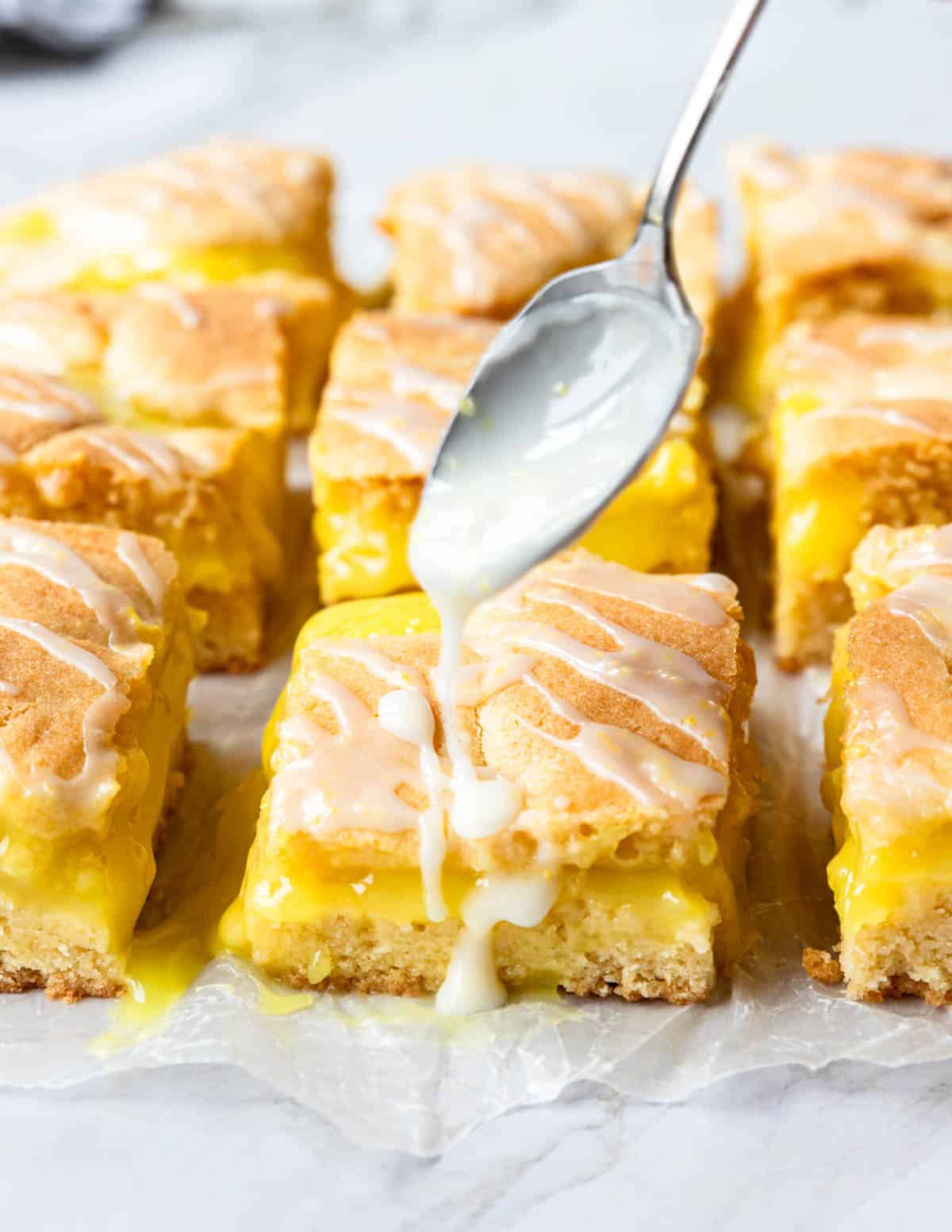lemon pie bars on a piece of parchment paper, being drizzled with lemon glaze