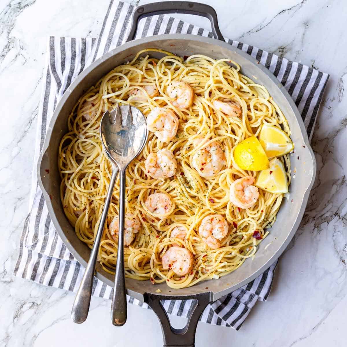 skillet of spaghetti with shrimp and a white wine garlic sauce, two serving spoons and lemon wedges set on top