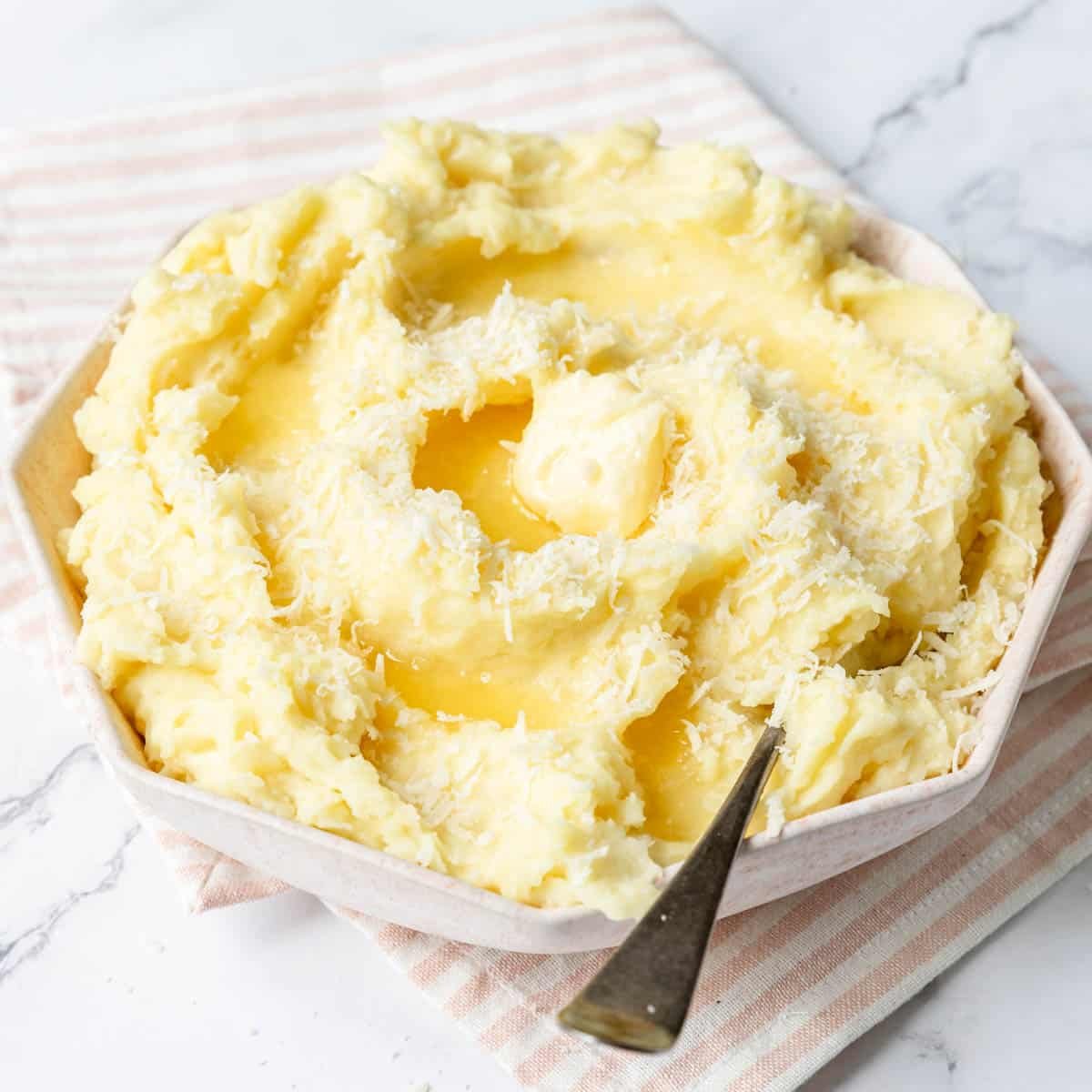 Garlic parmesan mashed potatoes served in a large serving bowl with grated parmesan and melted butter garnished on top with a serving spoon resting in the mixture.