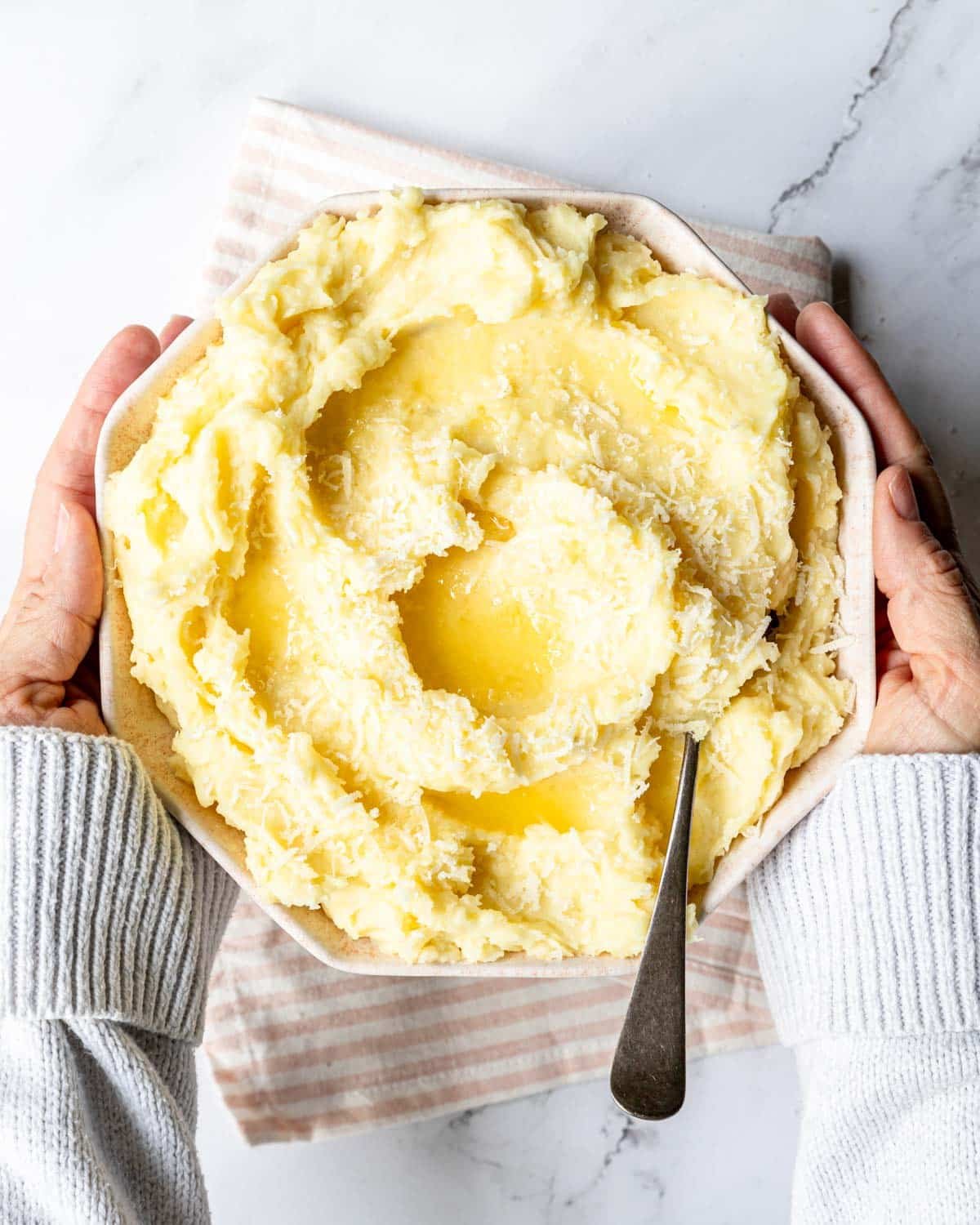 Hands picking up a large bowl of creamy garlic parmesan mashed potatoes with melted butter over the top and a serving spoon resting in it.