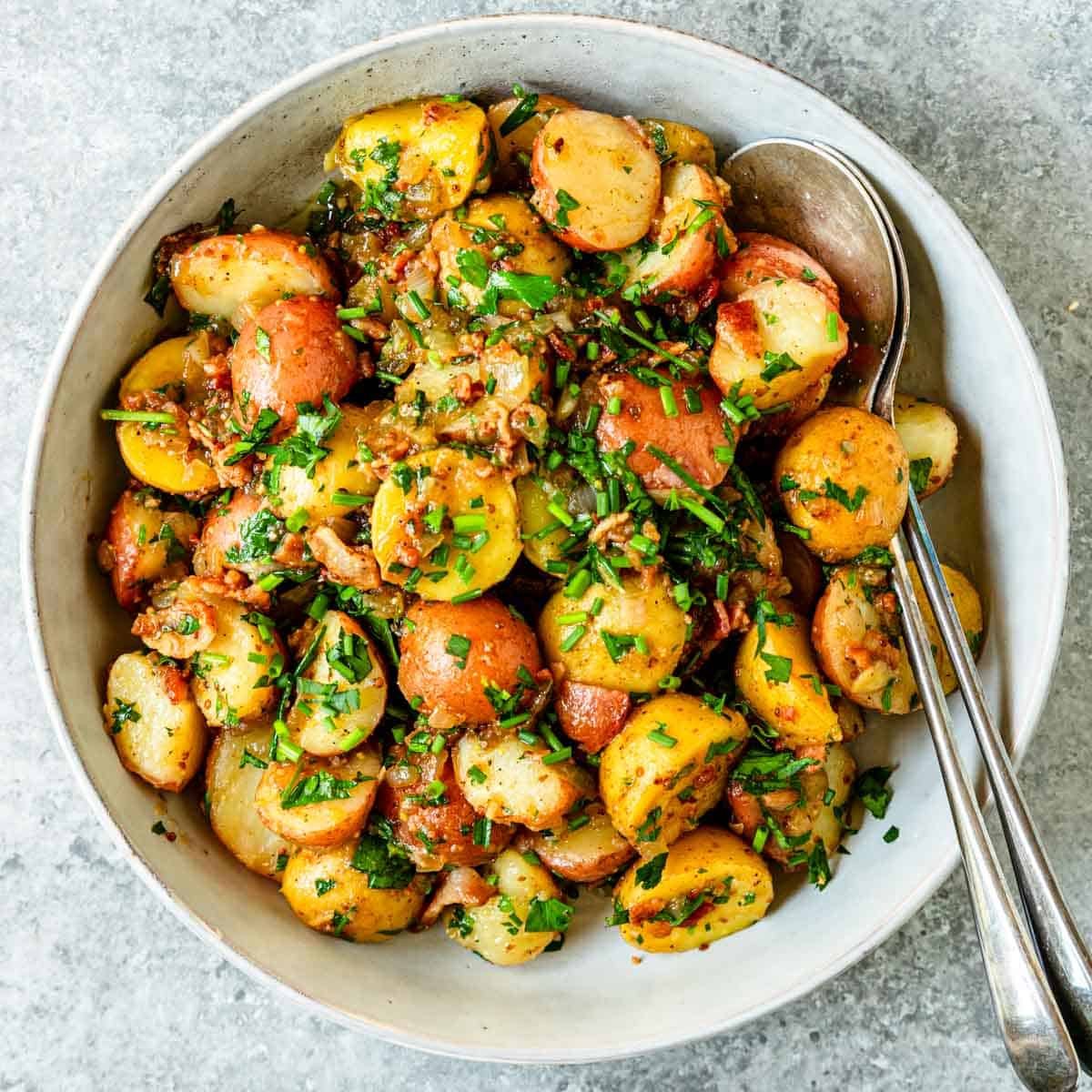 Top view of a large serving bowl that has chopped chives sprinkled over German potato salad with serving spoons resting inside.