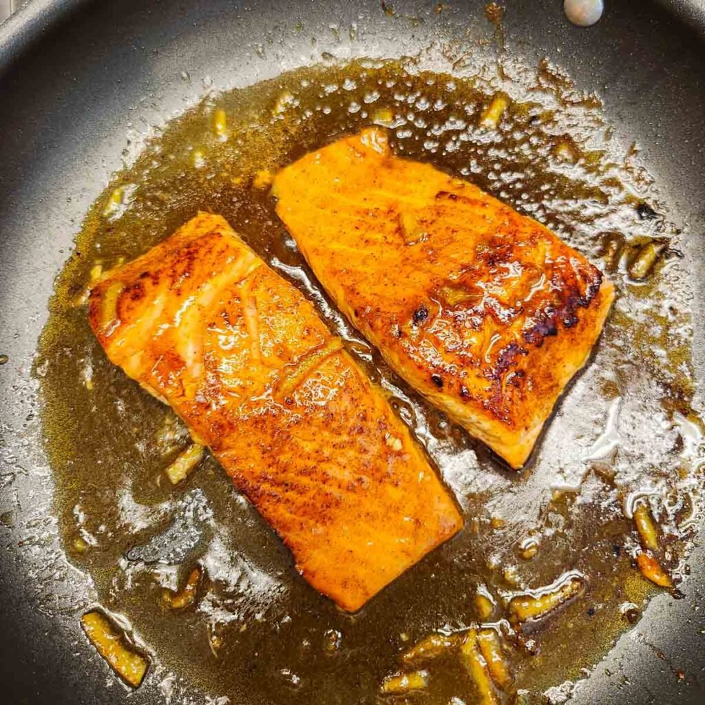 Seasoned salmon filets in a skillet with a glaze simmering around it.