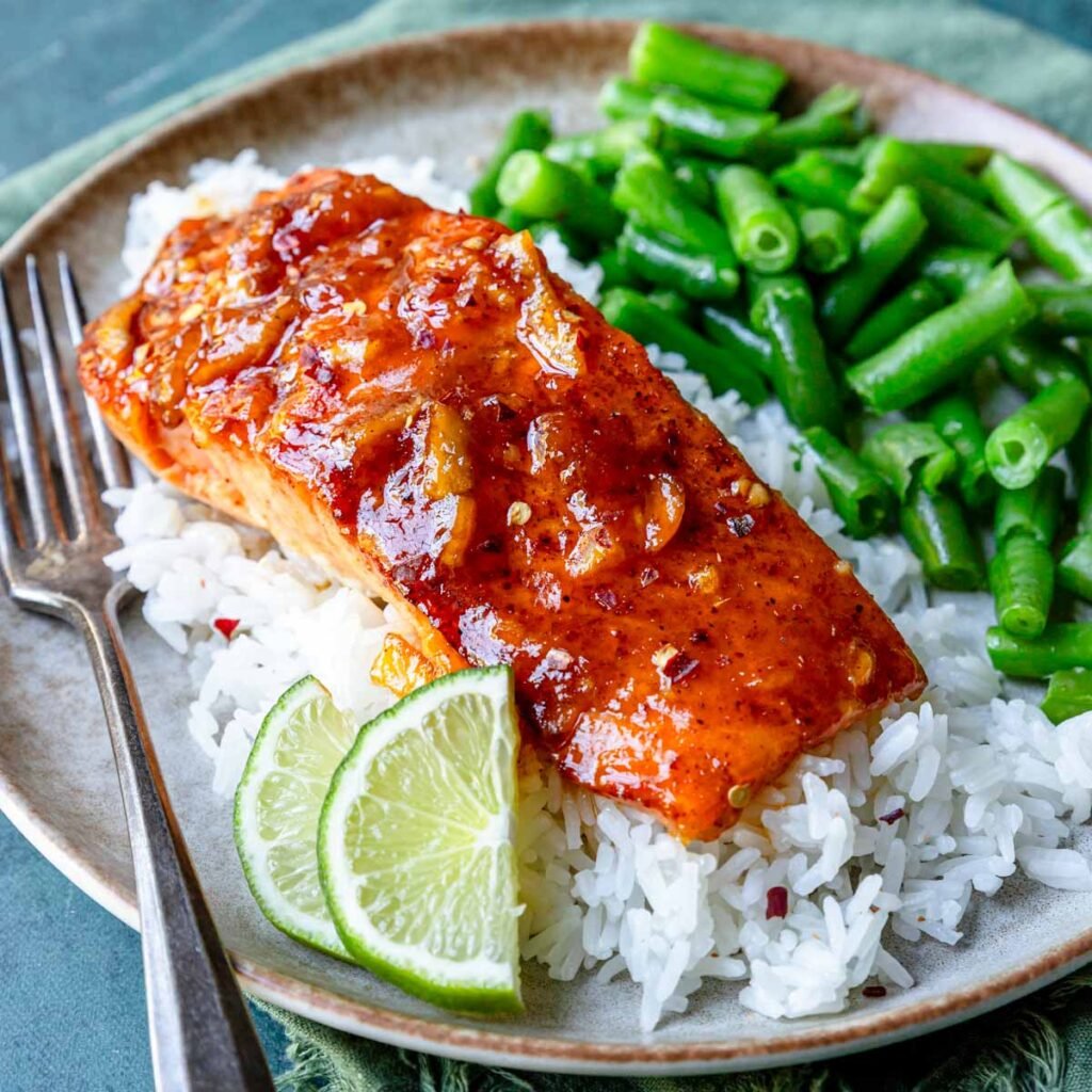 Plate of white rice topped with a glazed salmon filet with a side of green beans and two lime slices garnished on the side.