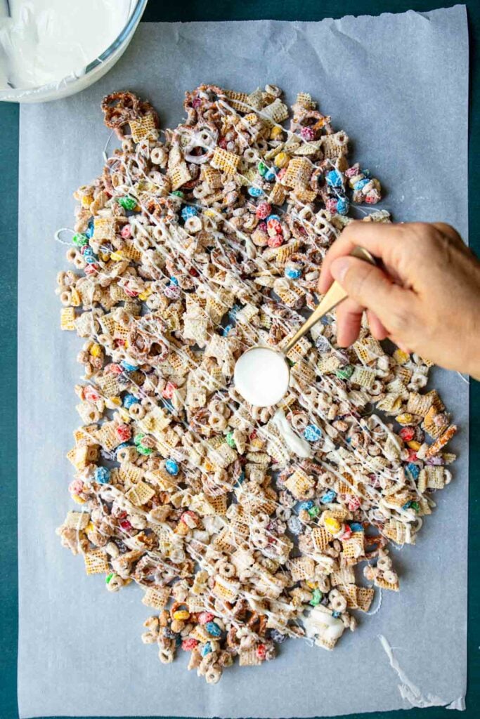 Spoon drizzling white chocolate over white chocolate chex mix that's been combined and drying on parchment paper.