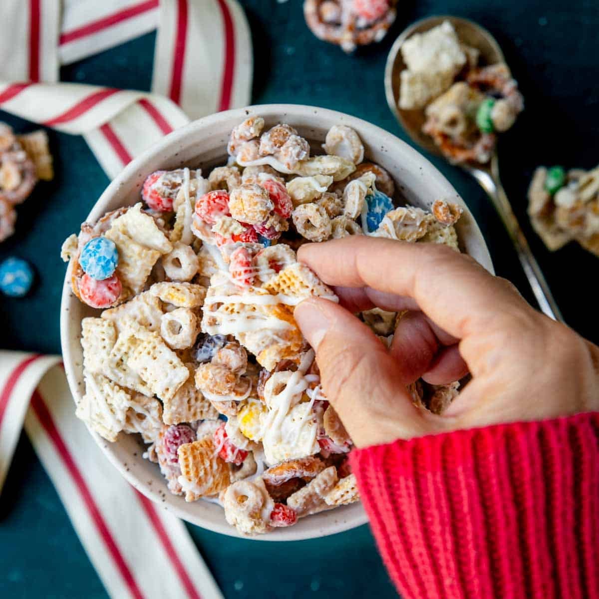 Fingers picking up a chunk of sweet chex mix from a bowl.