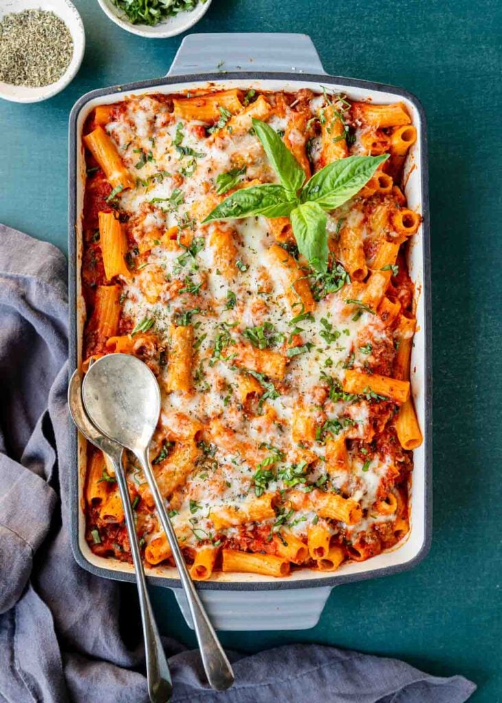 Top view of a freshly baked rigatoni pasta bake with a fresh sprig of basil on top and serving spoons resting on the side.