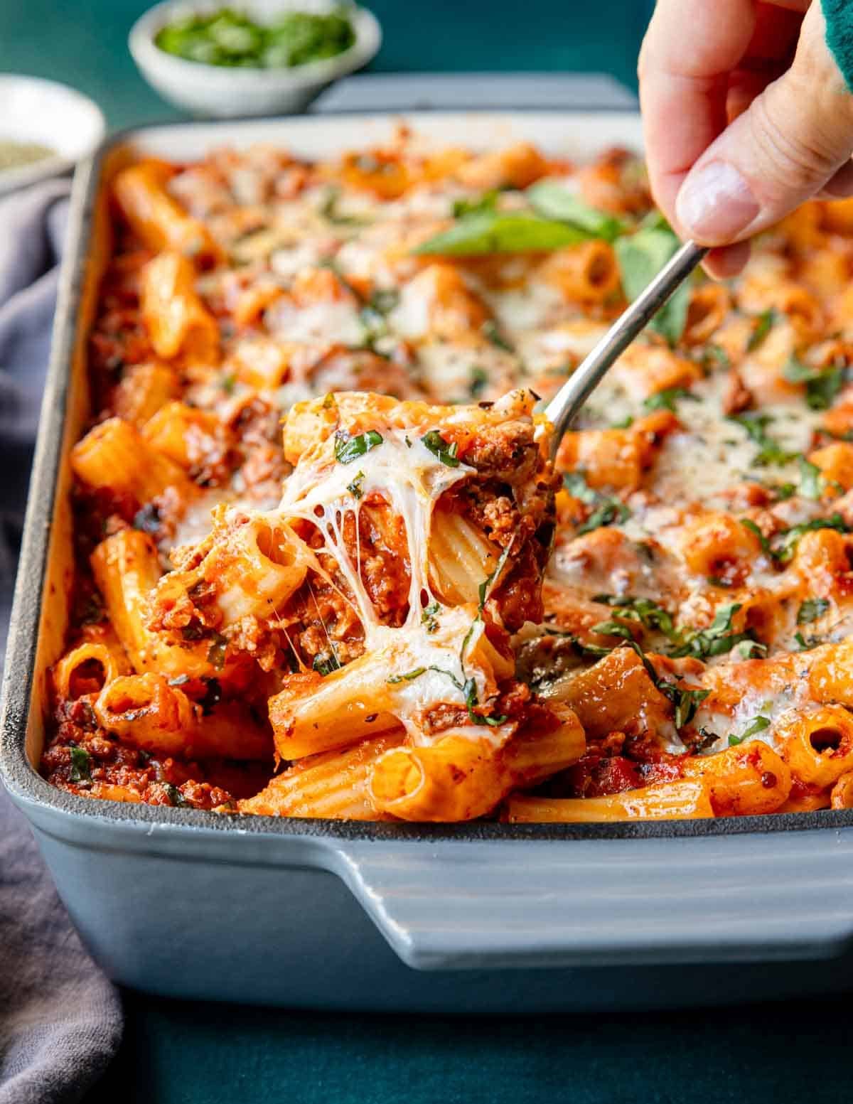 Hand scooping out a portion of baked rigatoni with melty cheese coming away from the pasta, from the baking dish.