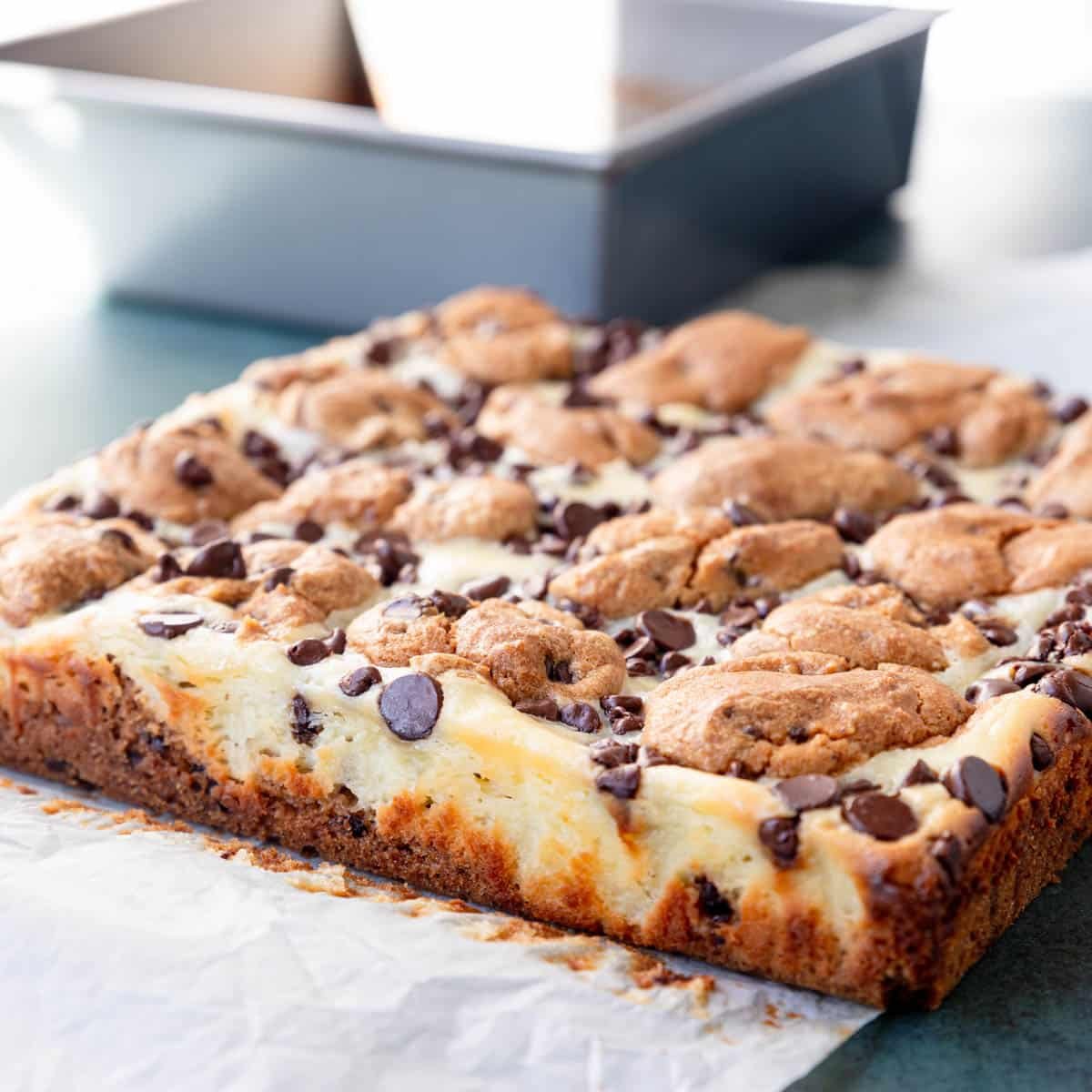 Side shot of chocolate chip cheesecake bars removed from the baking pan and sitting on parchment paper showing the golden crust and cheesecake filling with cookies baked into the top.