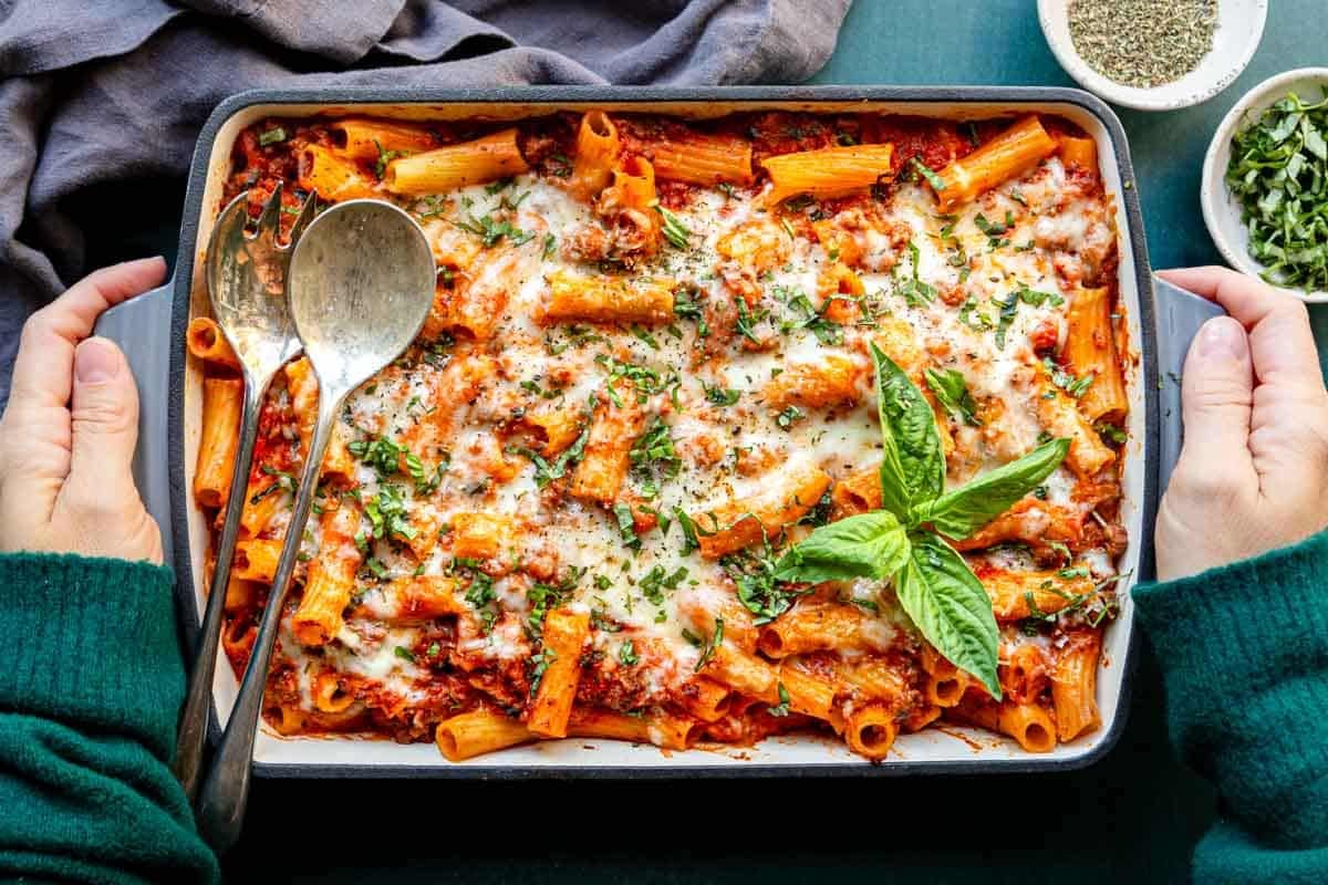 Hands placing a rigatoni bake on the table with serving spoons to share.