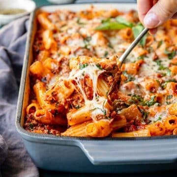 Hand scooping up a serving of baked rigatoni with a cheesy pull coming from the pasta.