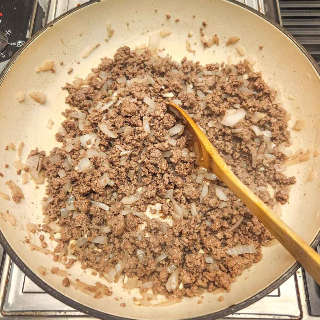 Browned ground beef and onions in a skillet with a wooden serving spoon stirring it.