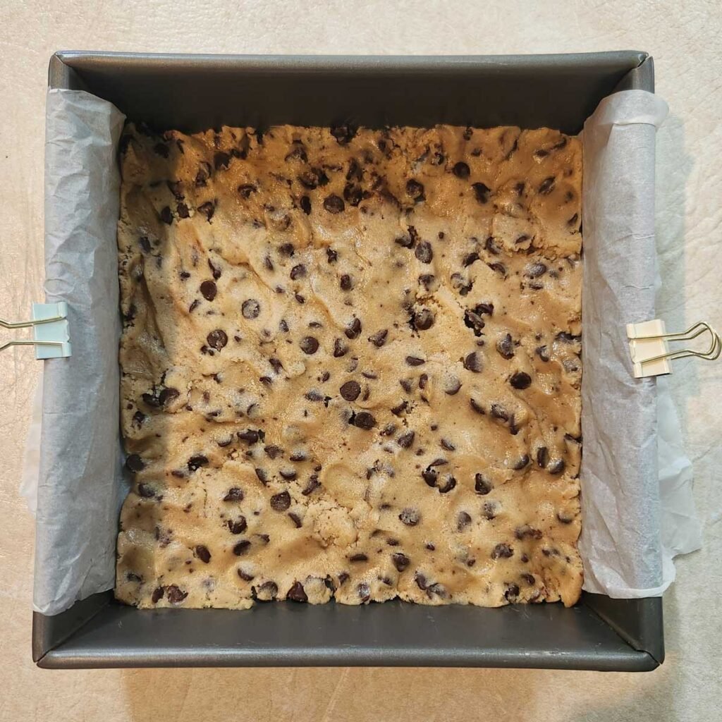 Raw chocolate chip cookie dough pressed over parchment paper in a baking dish.