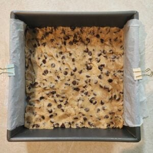 Raw chocolate chip cookie dough pressed over parchment paper in a baking dish.