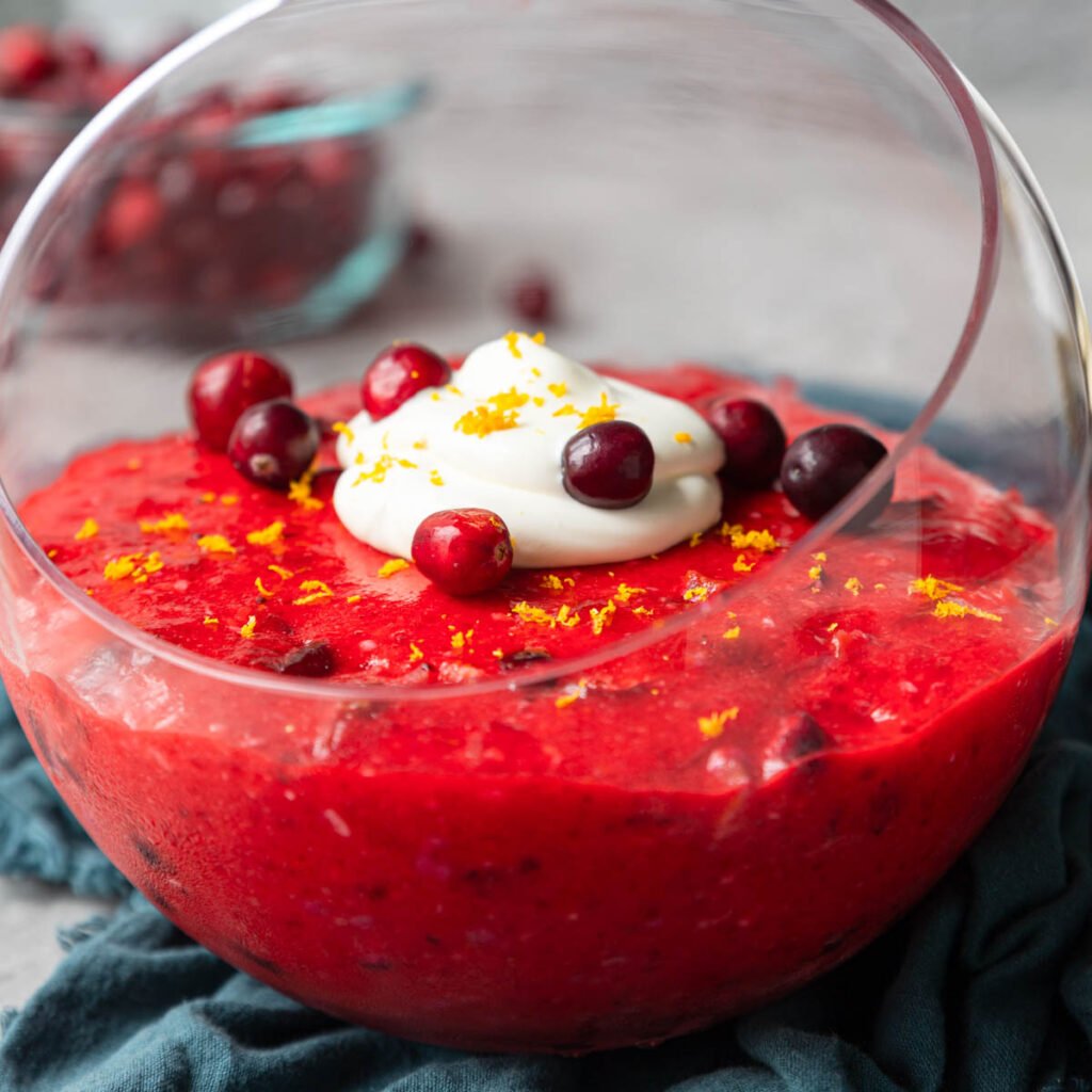 a glass bowl of cranberry jello salad with fresh cranberries and whipped cream on top