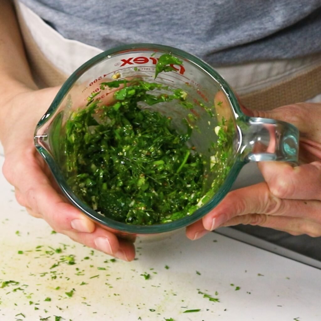 Hand holding up fresh herbs mixed with seasoning and oil in a glass measuring cup.