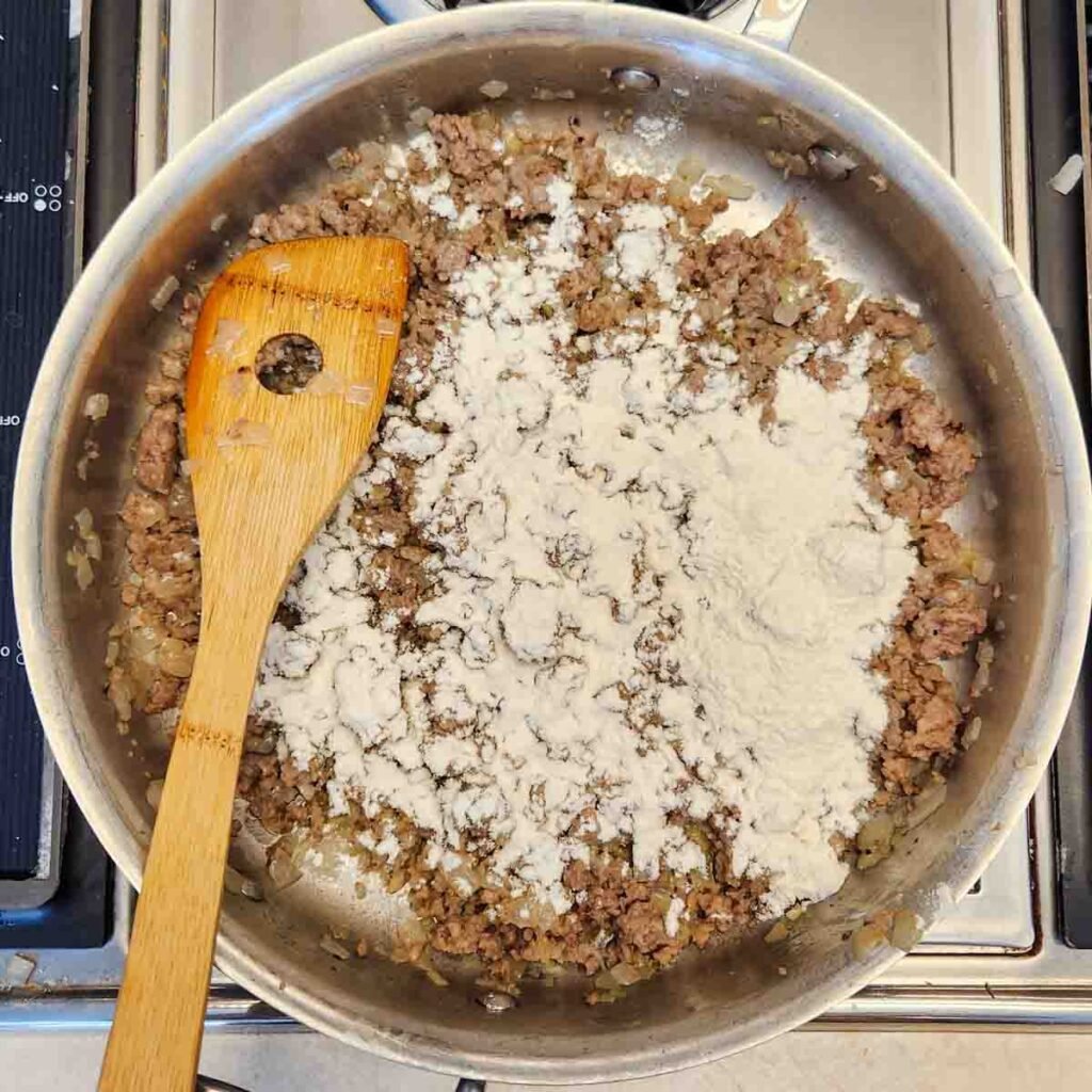 Flour sprinkled over browned ground sausage in a skillet.