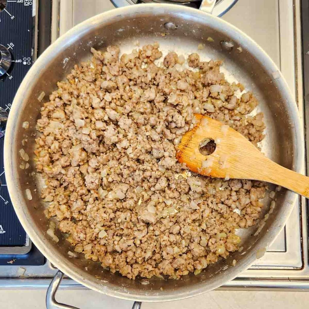 Ground sausage browned in a skillet with a wooden spoon resting in the mixture.