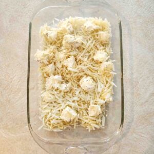 Shredded hashbrowns in a glass baking pan topped with cubes of cream cheese topped with salt and pepper.