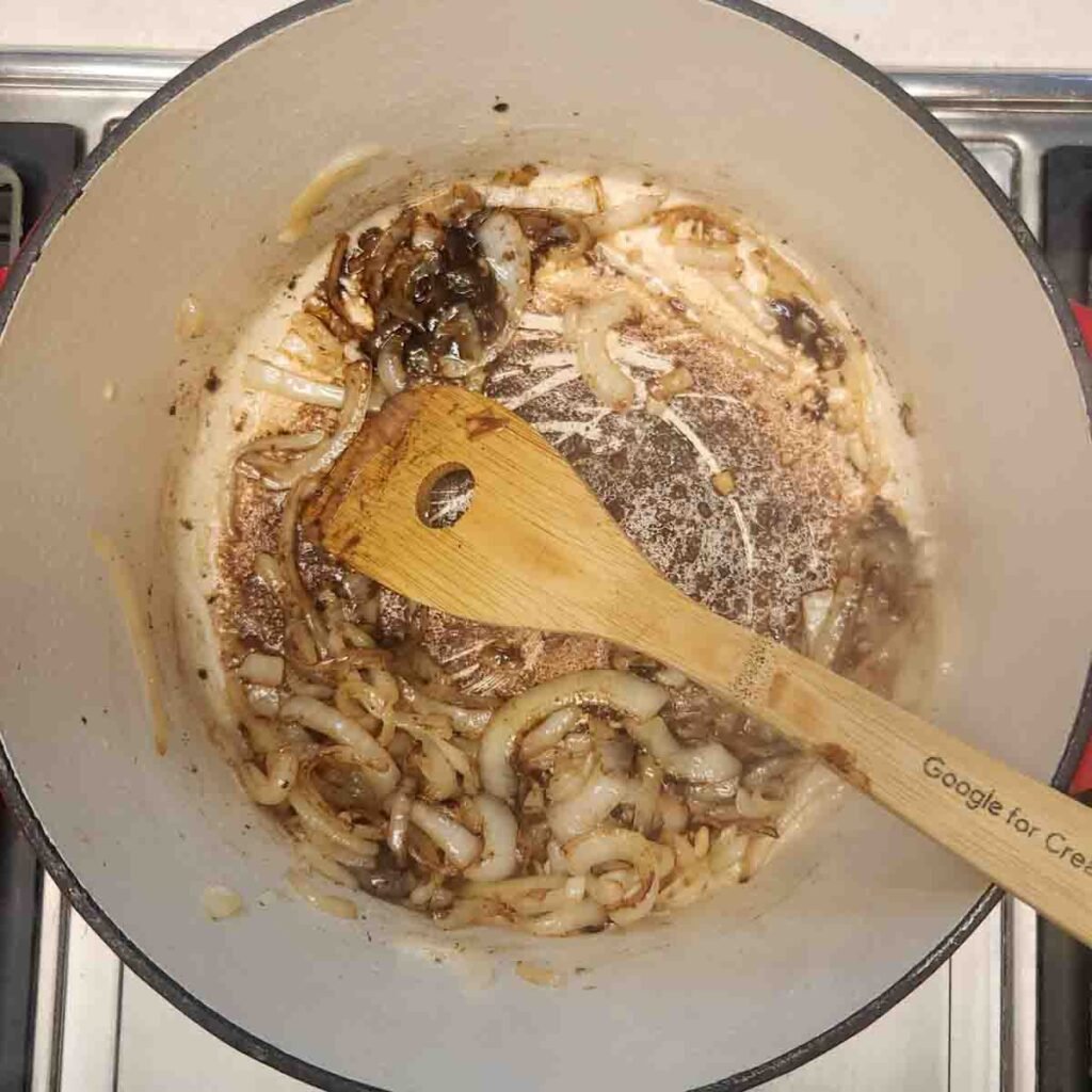 Wooden spoon resting in a Dutch oven stirring sliced onions sauteeing.