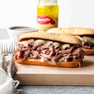 Two French dip sandwiches topped with melted cheese on a toasted sub bun next to a bowl of au jus for dipping.