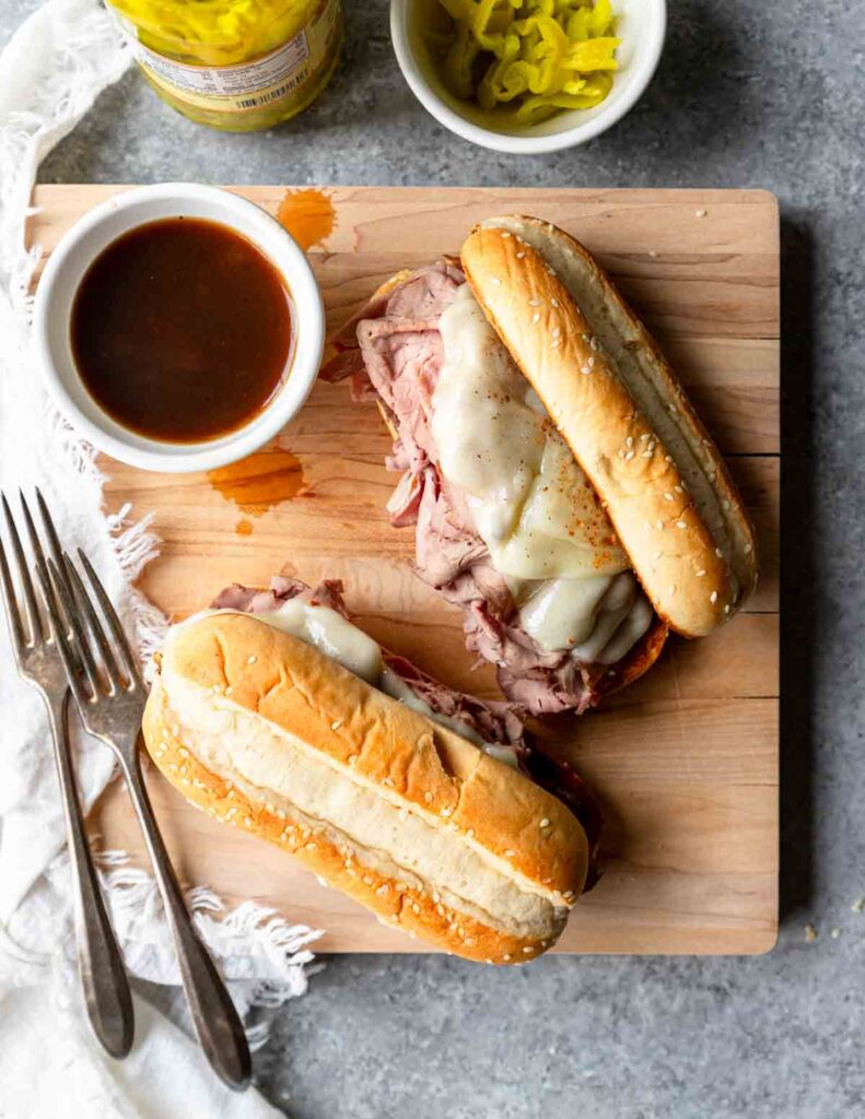 Top view of hot French dip sandwiches on a wooden cutting boar with a small bowl of au jus next to them.