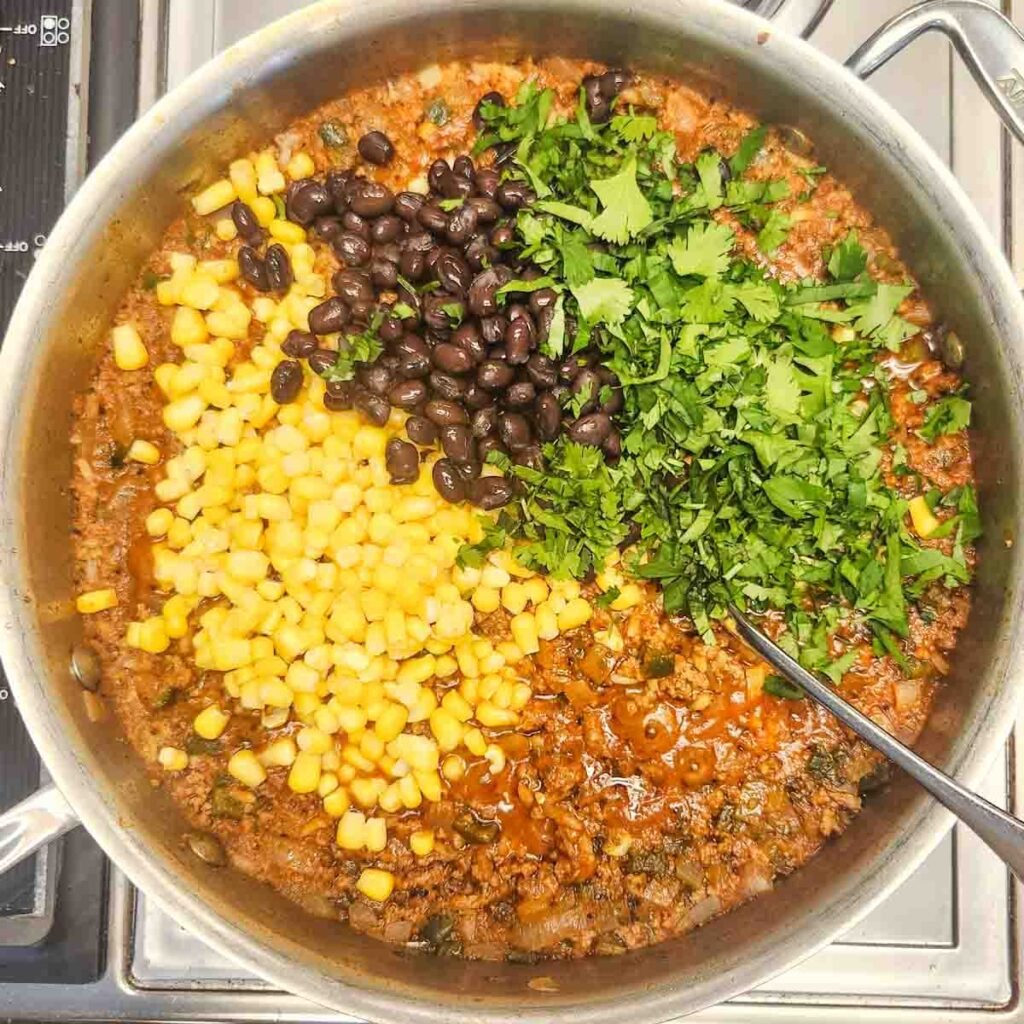 Beans and corn with chopped cilantro added to the beef mixture in a skillet.