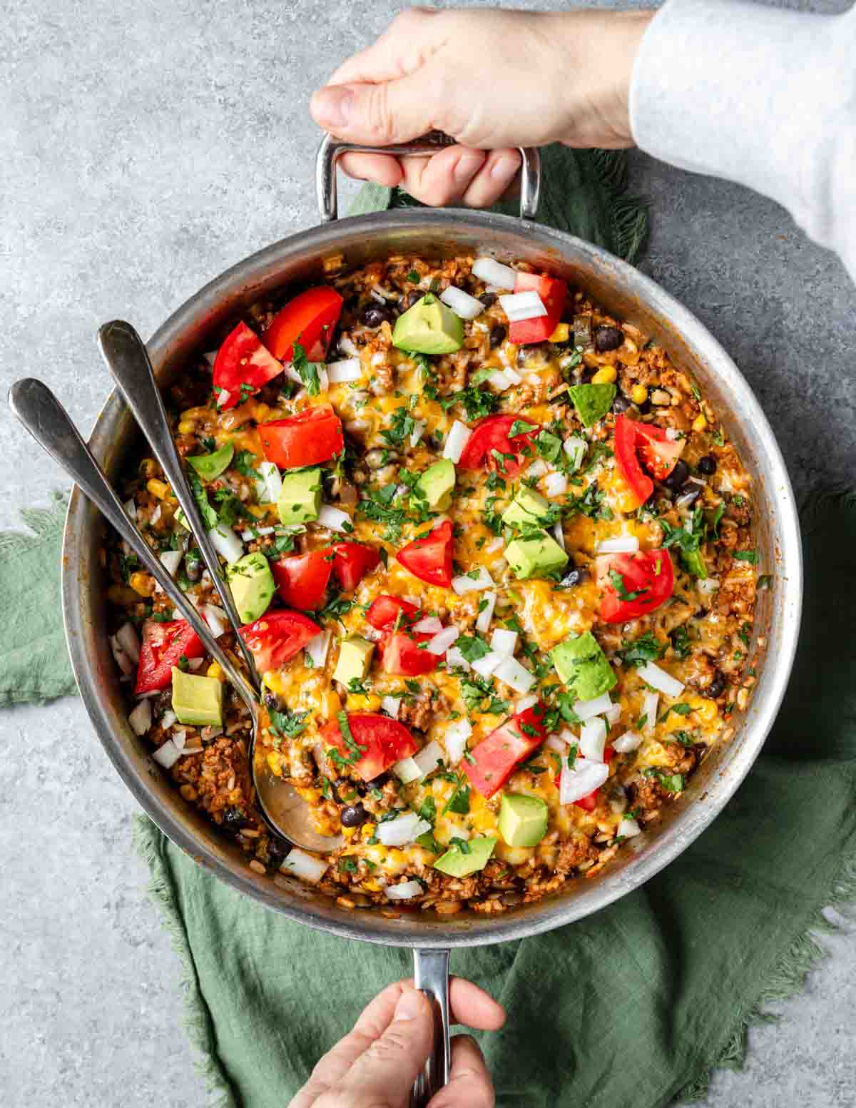 Hands setting a skillet on a green cloth with a hot taco and beef skillet served, garnished with cilantro, avocado and tomatoes over top.