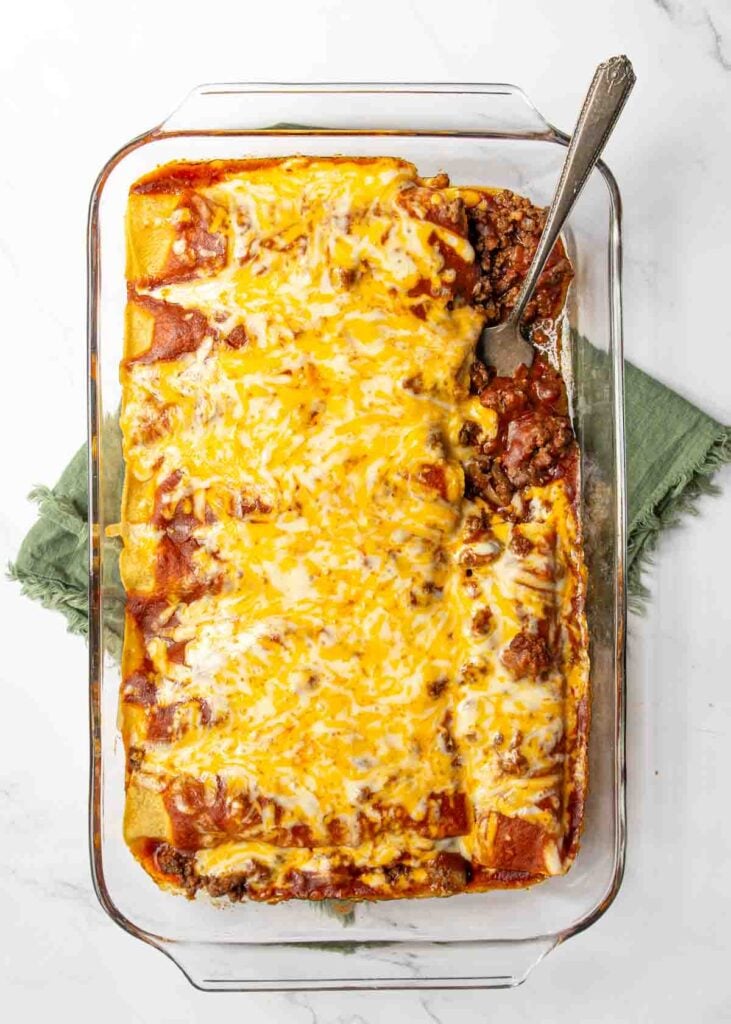 Fork resting in a freshly baked pan of beef enchiladas with melted cheese coating the enchilada sauce.