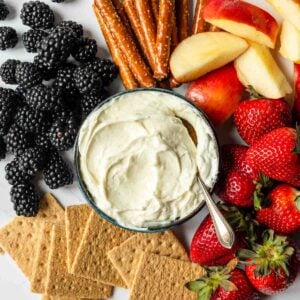 Fluff fruit dip in a bowl with a spoon resting in the mixture, with fruit, crackers and pretzels surrounding it.