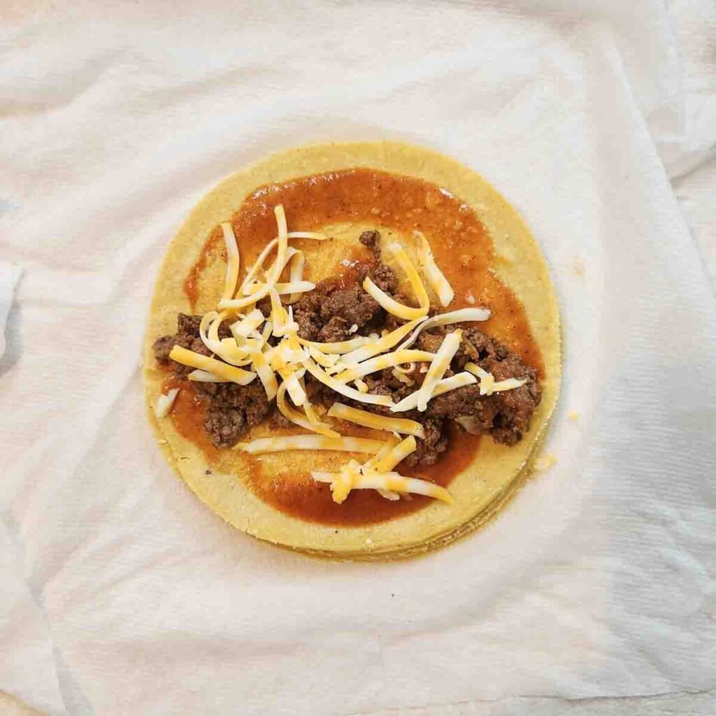 Red sauce, ground beef and shredded cheese topping a yellow corn tortilla.