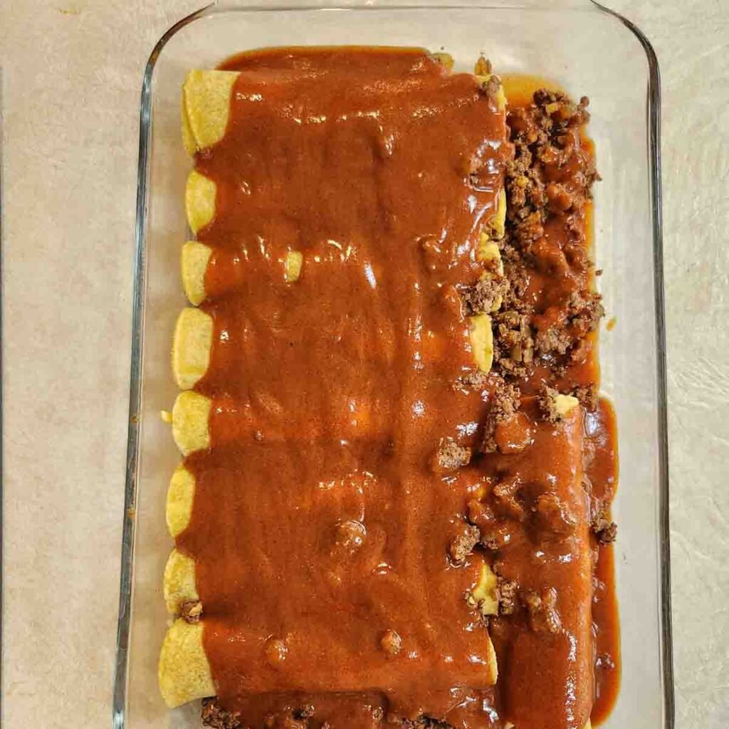 Red enchilada sauce covering corn tortilla wrapped ground beef enchiladas in a glass baking dish.