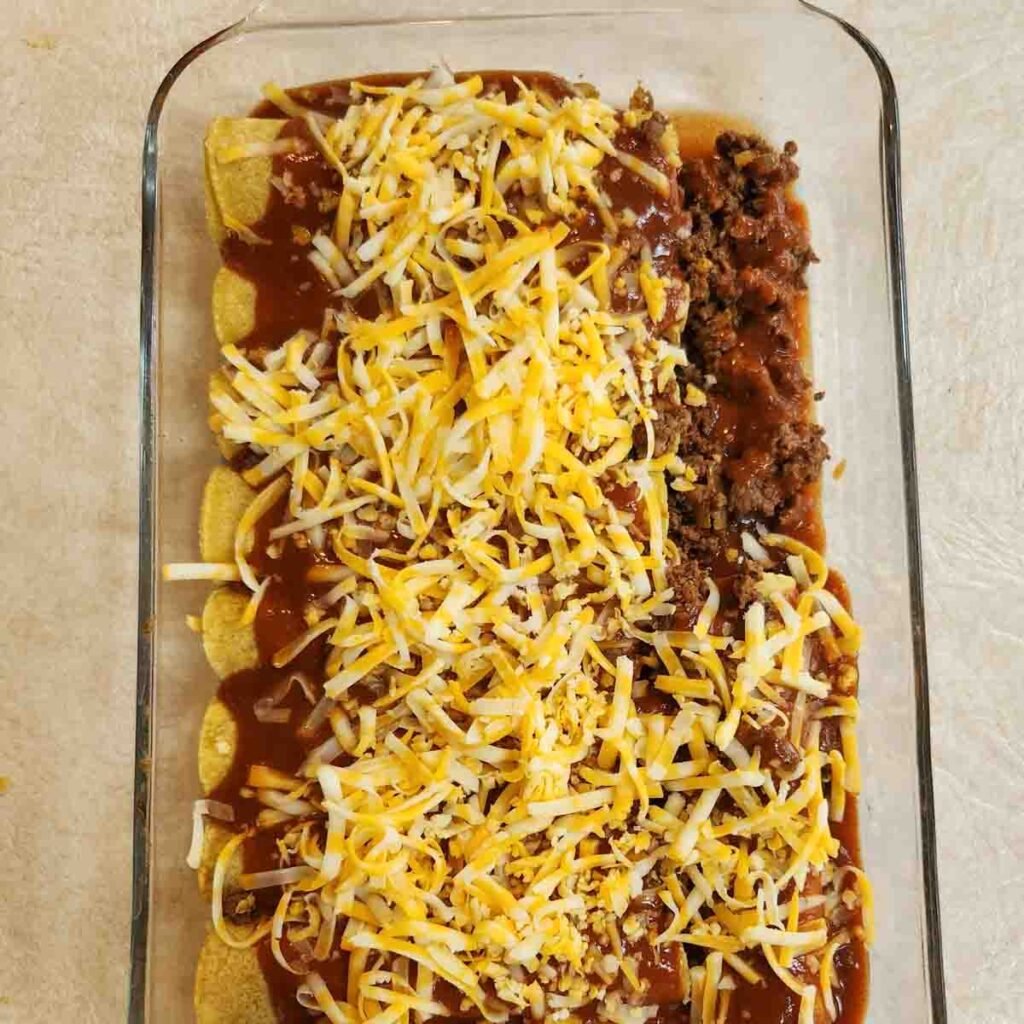 Shredded cheese sprinkled over ground beef enchiladas wrapped in corn tortillas in a baking dish.
