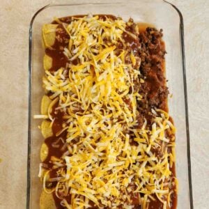 Shredded cheese sprinkled over ground beef enchiladas wrapped in corn tortillas in a baking dish.