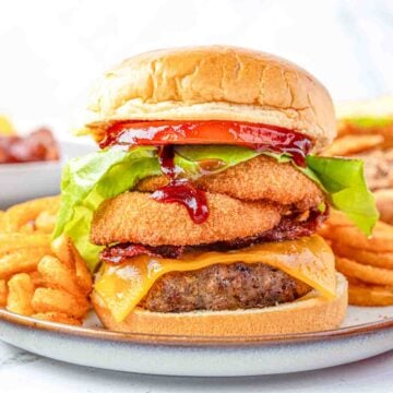 Dinner plate serving a barbecue bacon cheeseburger with onion rings and curly fries on the side.