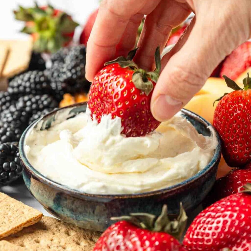 A hand dipping a strawberry into marshmallow fruit dip 