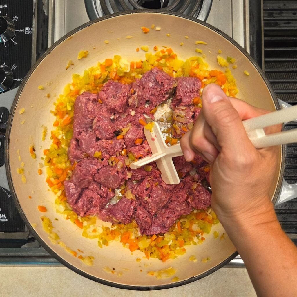Hand using a meat chopper to cook ground beef in a pan with a satueed sofrito.