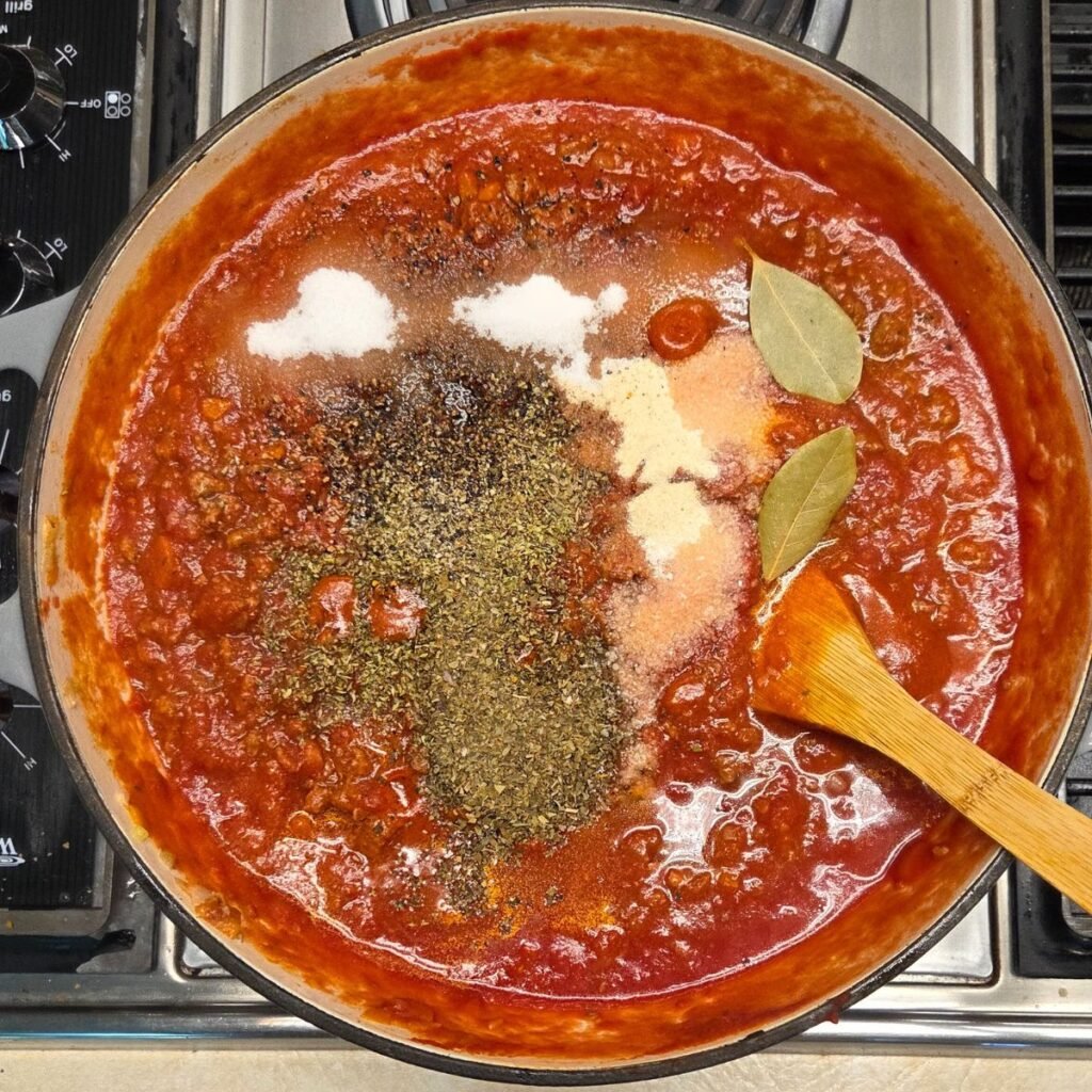 Seasonings and bay leaves measured out and added to sauce in a pan.