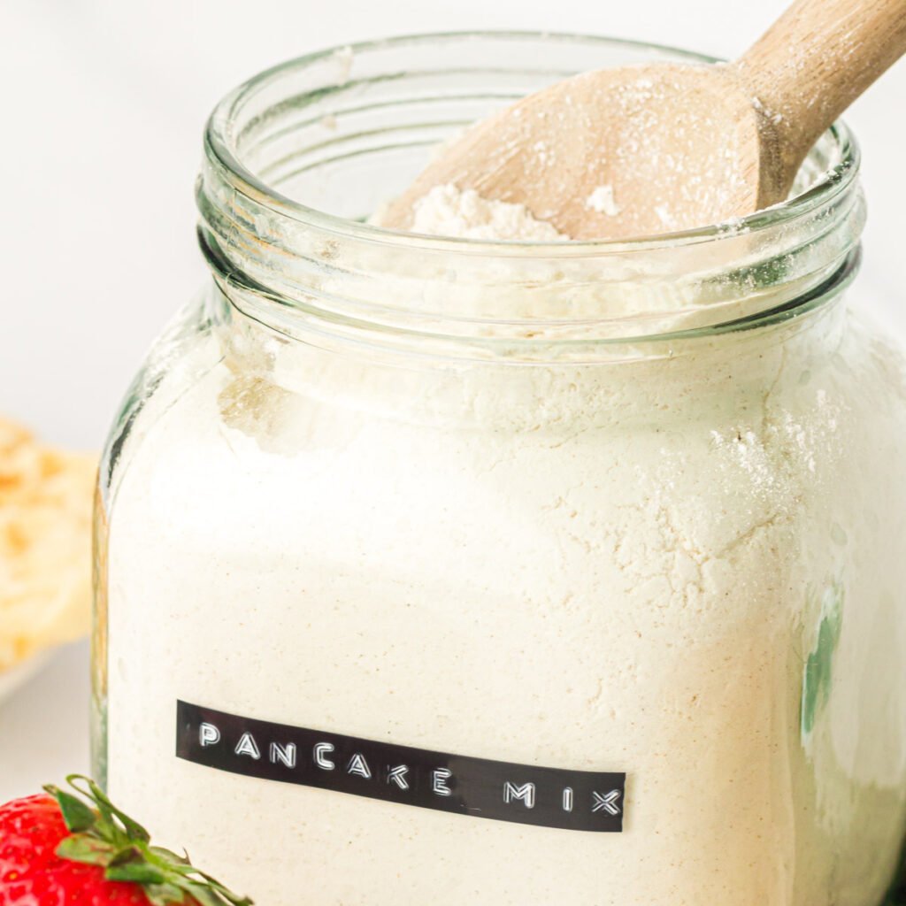 Wooden spoon dipping into a glass jar of dry pancake mix.