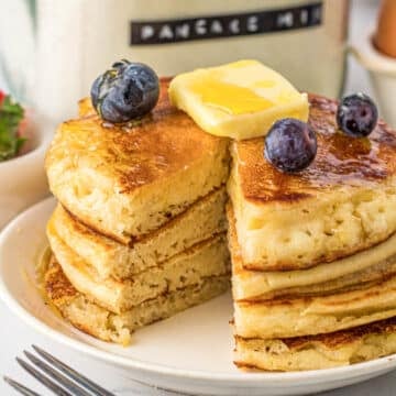 Stack of golden brown pancakes with a pat of butter melting on top with fresh blueberries garnished over the top and a bite of pancakes cut out.