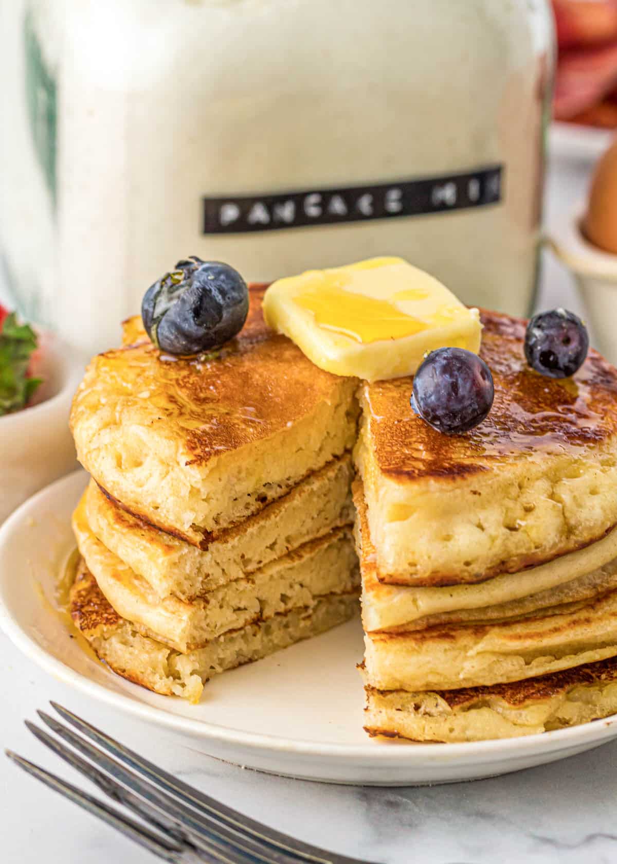 Dry pancake mix in a glass jar next to a plate of golden brown pancakes with a bite cut out from the syrup drenched breakfast.