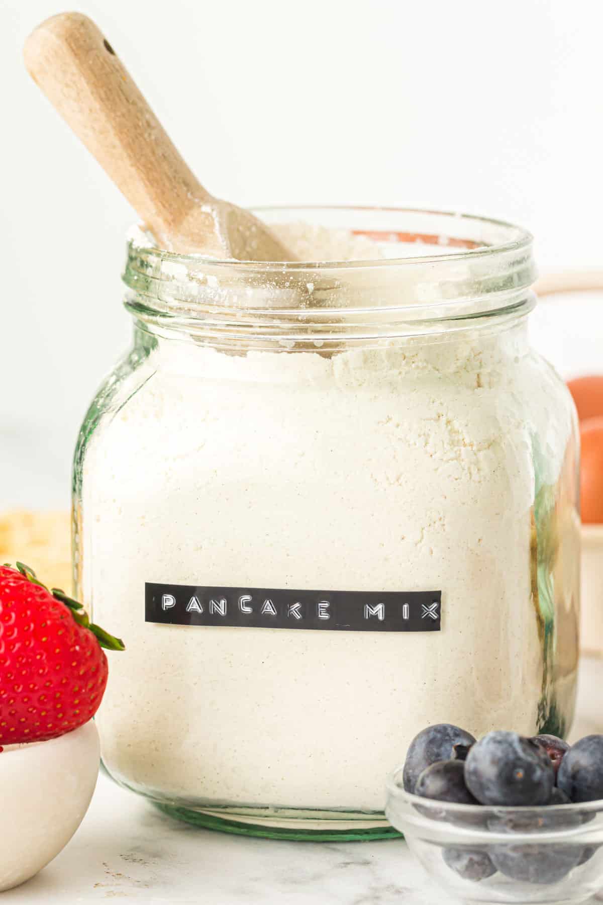 A bowl of fresh blueberries in front of a glass jar of dry pancake mix ready to use to prepare homemade pancakes.