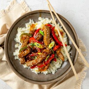 Steak stir fry served over a bed of white rice with chopsticks resting on the edge.