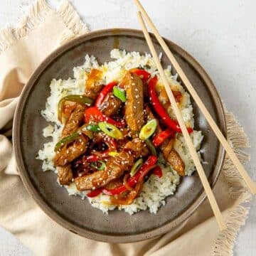 Steak stir fry served over a bed of white rice with chopsticks resting on the edge.