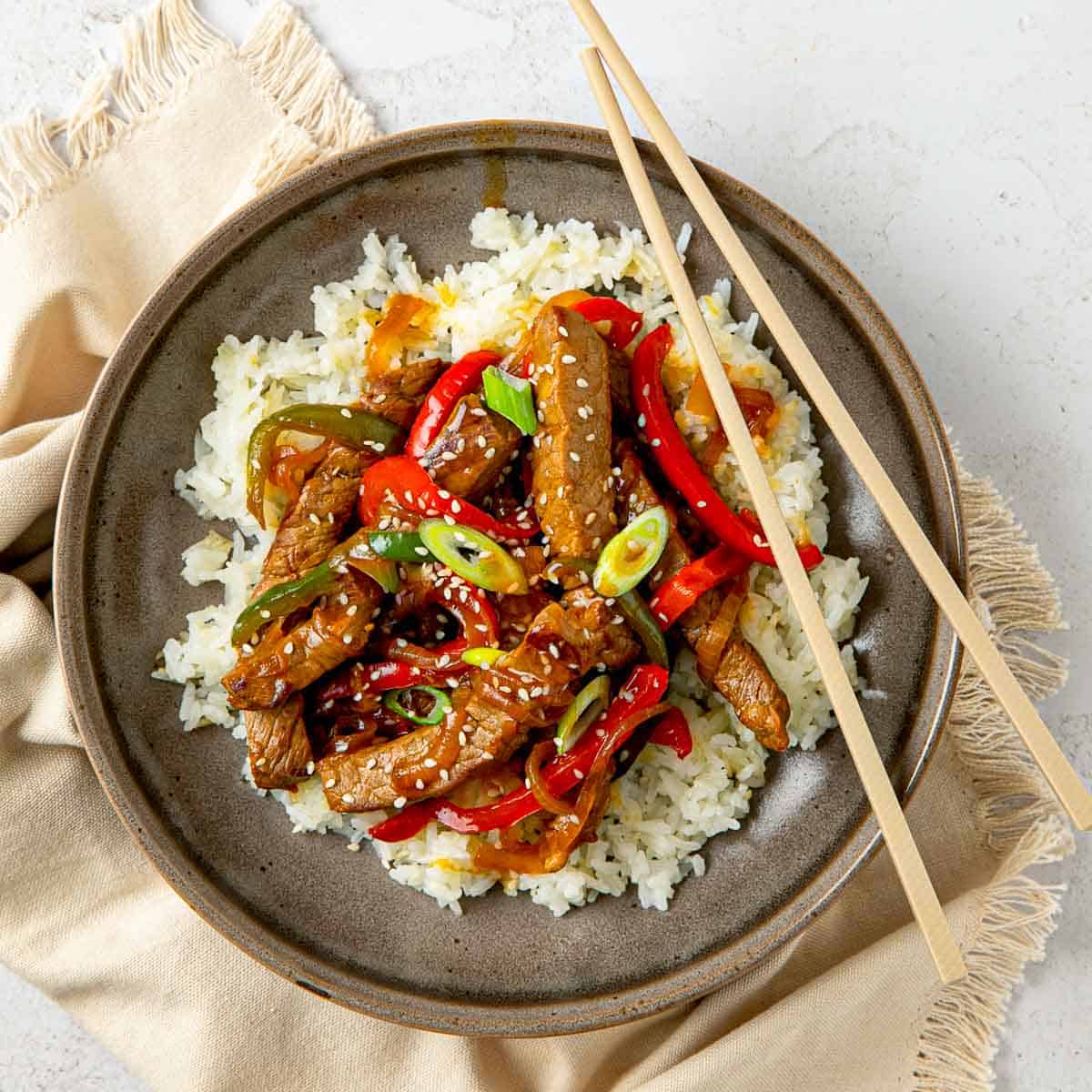 Steak stir fry served over a bed of white rice with chopsticks resting on the edge.