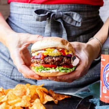 A woman holding a grilled steak house burger on a bun with chips on a plate and a soda to the side