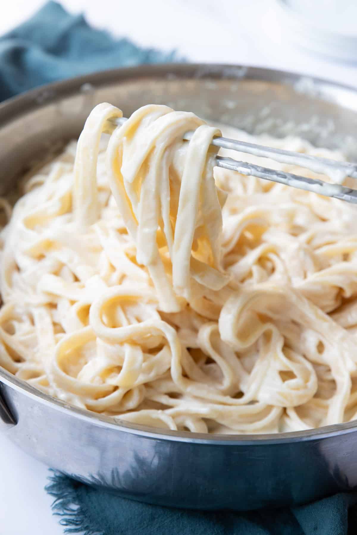 Homemade creamy alfredo sauce coating linguine noodles being lifted out of a pan with tongs.